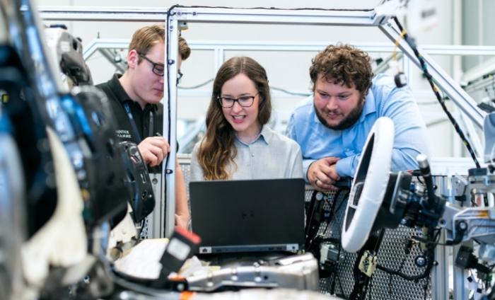 Photo of two men and one woman gathered around a laptop in a car manufacturing plant. 